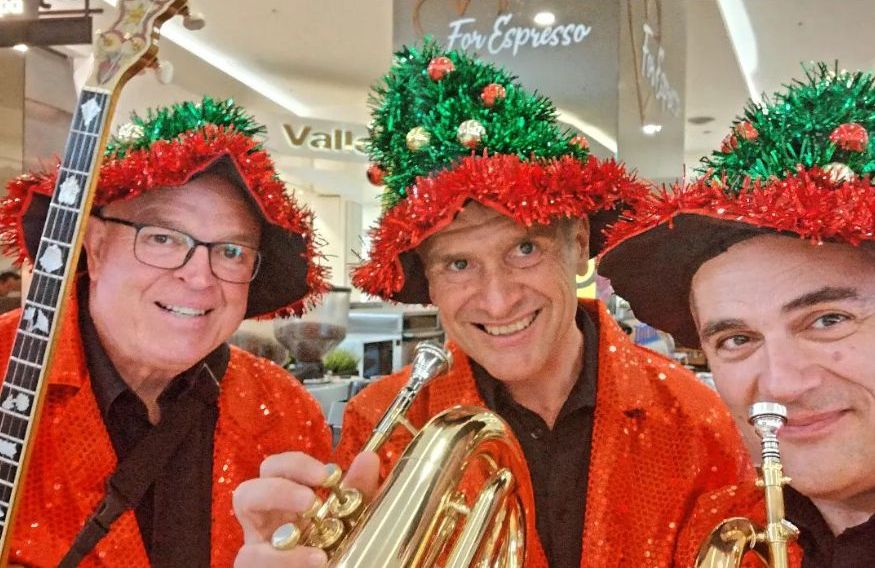 Three musicians in Christmas outfits smile for the camera in a shopping centre.